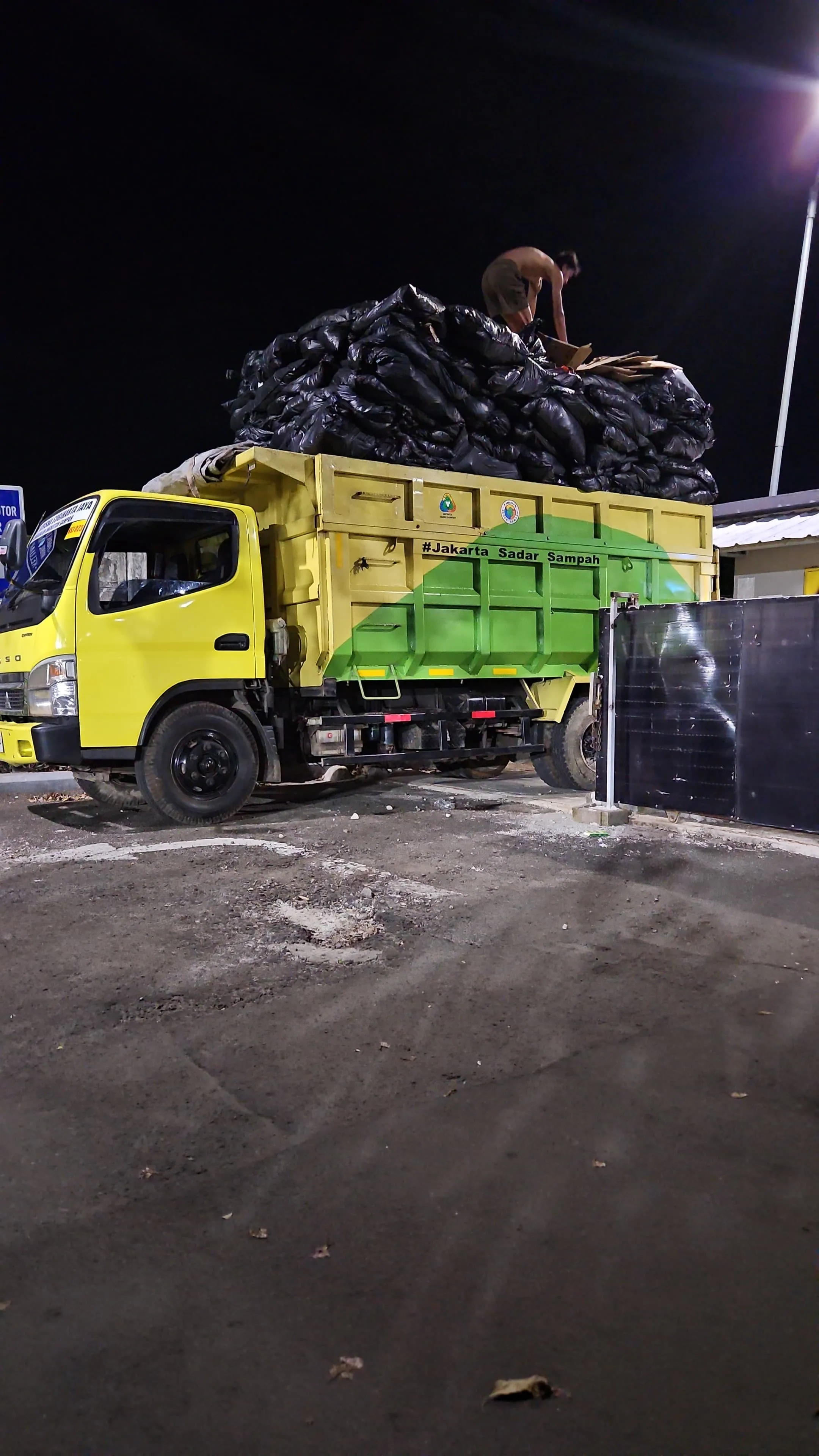 Dump truck kuning dan hijau membawa muatan sampah dalam kantong tertutup.